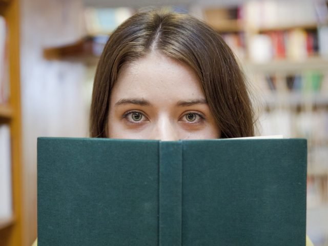 chica-adolescente-que-cubre-la-cara-con-el-libro-en-la-biblioteca